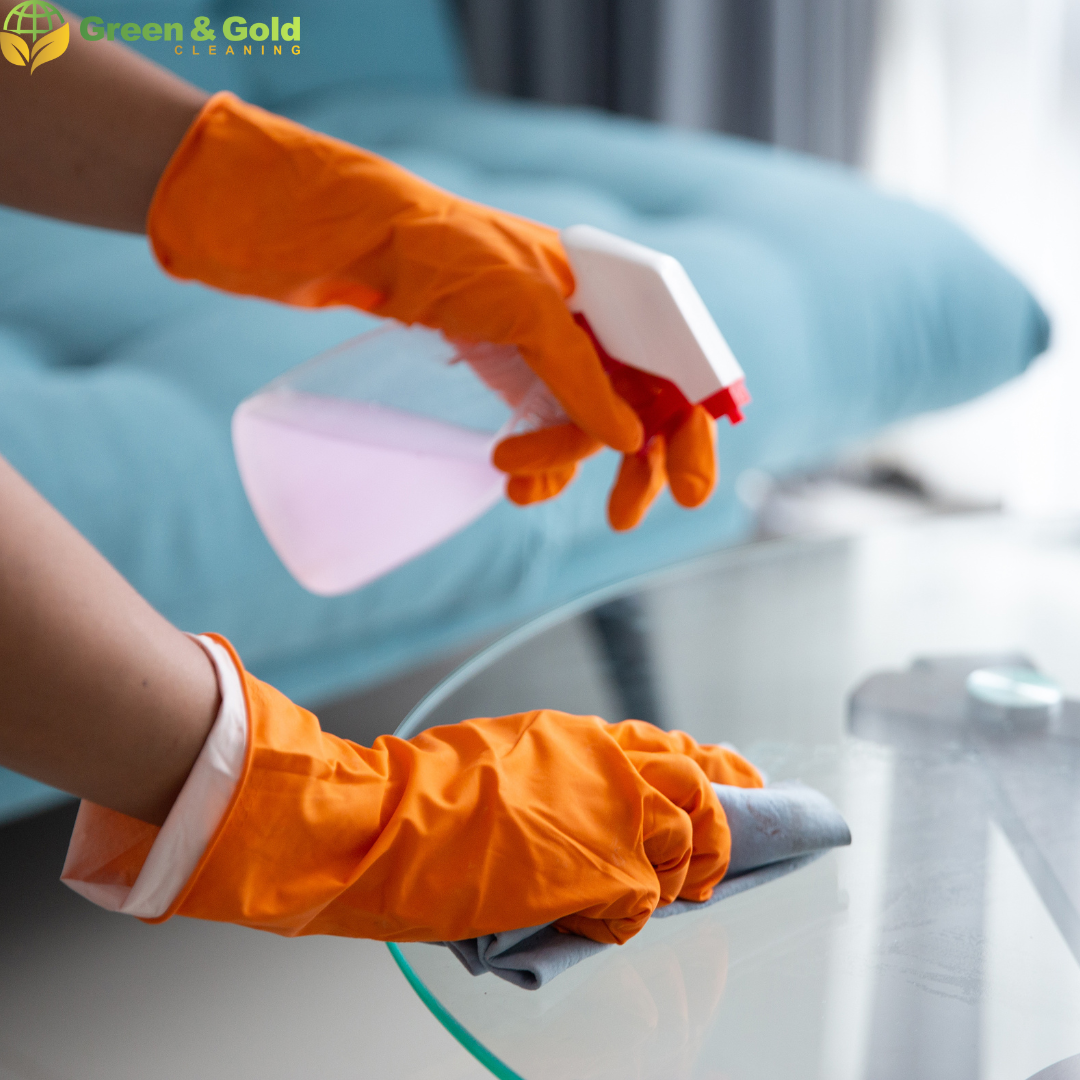 Close-up of a person's hand wearing orange rubber gloves, cleaning a glass table with a grey cloth and spraying a pink cleaning solution from a white spray bottle, with a blurred sofa in the background.