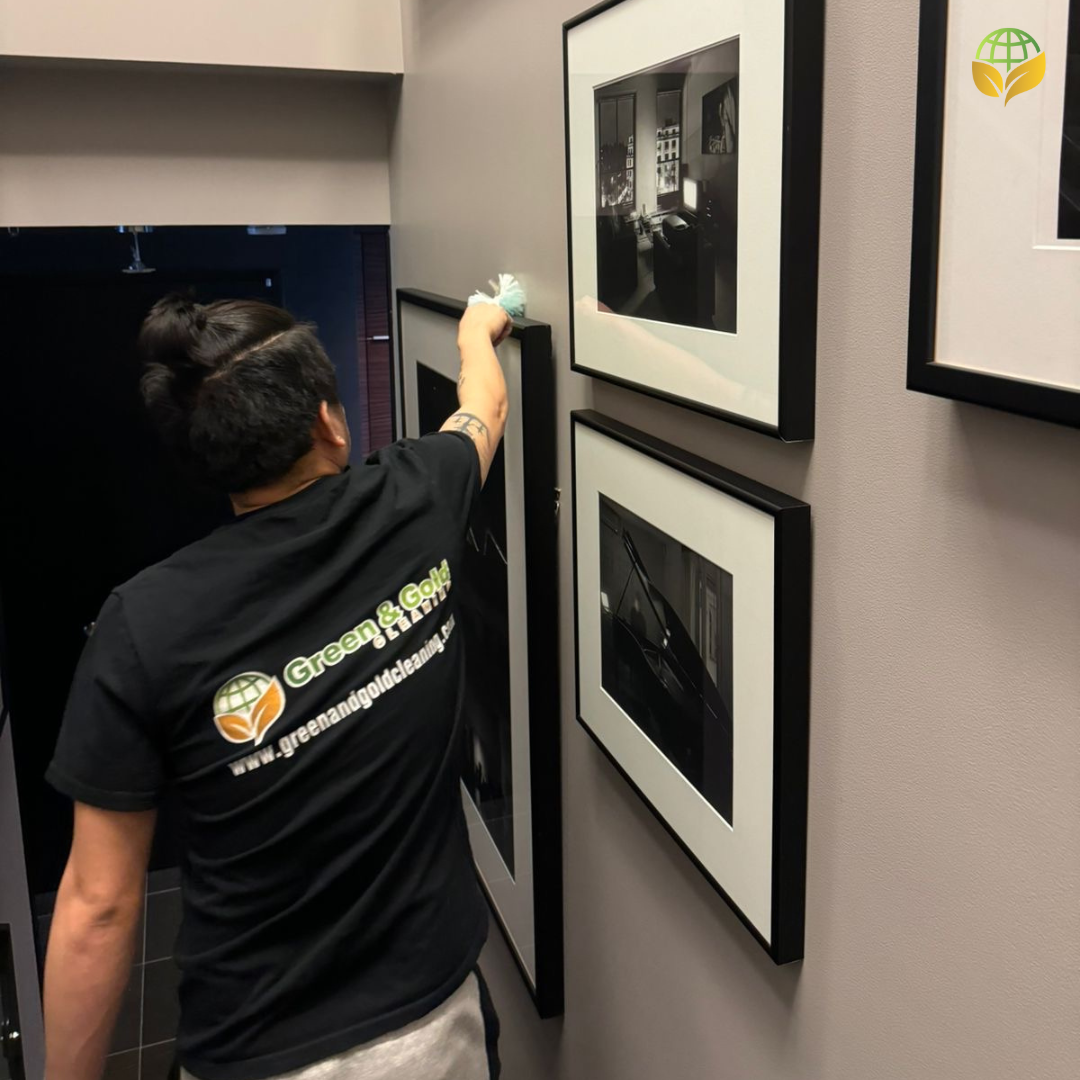 A person in a black shirt with the "Green & Gold Cleaning" logo cleaning a picture frame, demonstrating professional cleaning services' attention to detail.